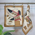 Two vintage Egyptian-style quilted cloth hangings from gold-colored hooks on a metal rod. The napkin on the left is fully displayed, showing a figure with a feathered headdress in beige, brown, black, red, and white.