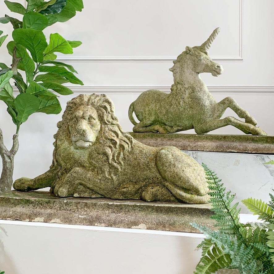 A pair of stone garden sculptures, featuring a detailed lion and a unicorn, displayed on a stone shelf with green plants in a home setting.
