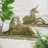 A pair of stone garden sculptures, featuring a detailed lion and a unicorn, displayed on a stone shelf with green plants in a home setting.