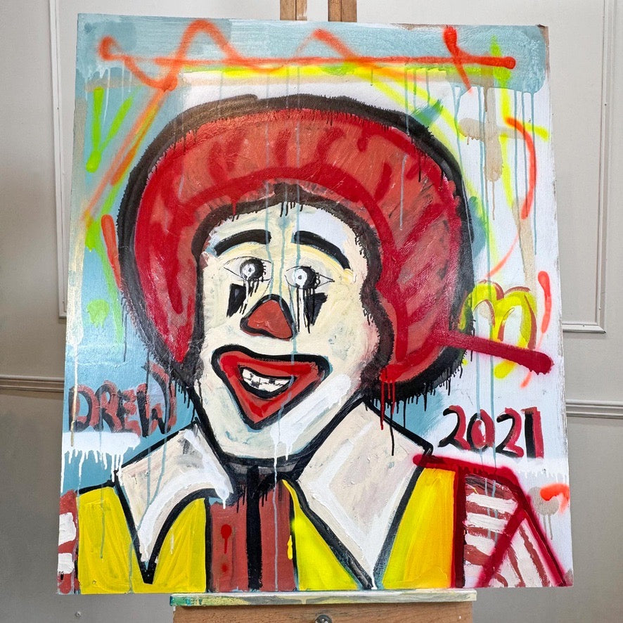 A colorful portrait painting of a clown-like figure with a large red hat, white face, red lips, and yellow clothing, shown on an easel against an indoor wall background. The painting includes the text 'DREW' and '2021'.