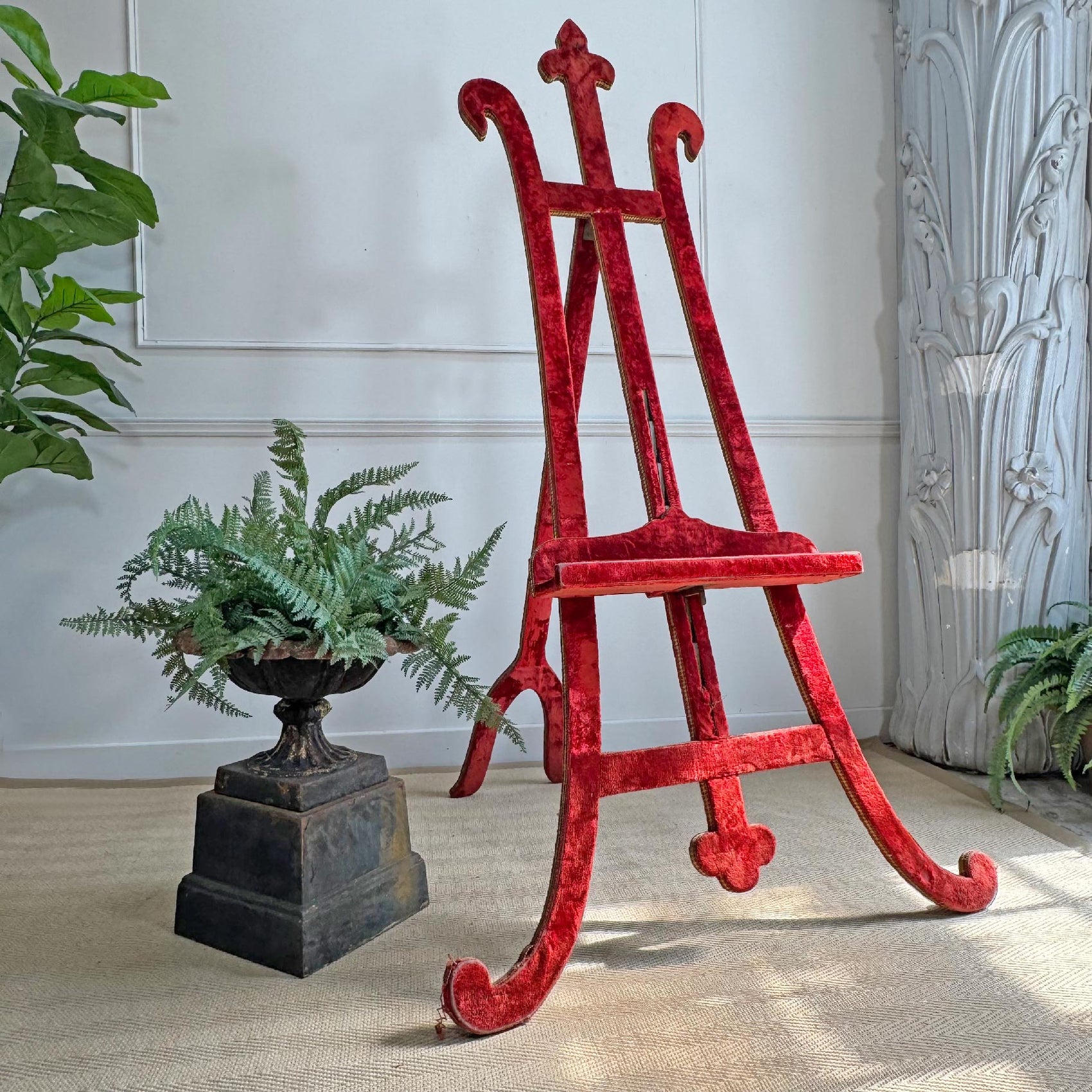 An antique red velvet covered wooden easel with curved legs and a decorative finial on the top, styled in a room with potted ferns and a textured wall.