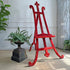 An antique red velvet covered wooden easel with curved legs and a decorative finial on the top, styled in a room with potted ferns and a textured wall.