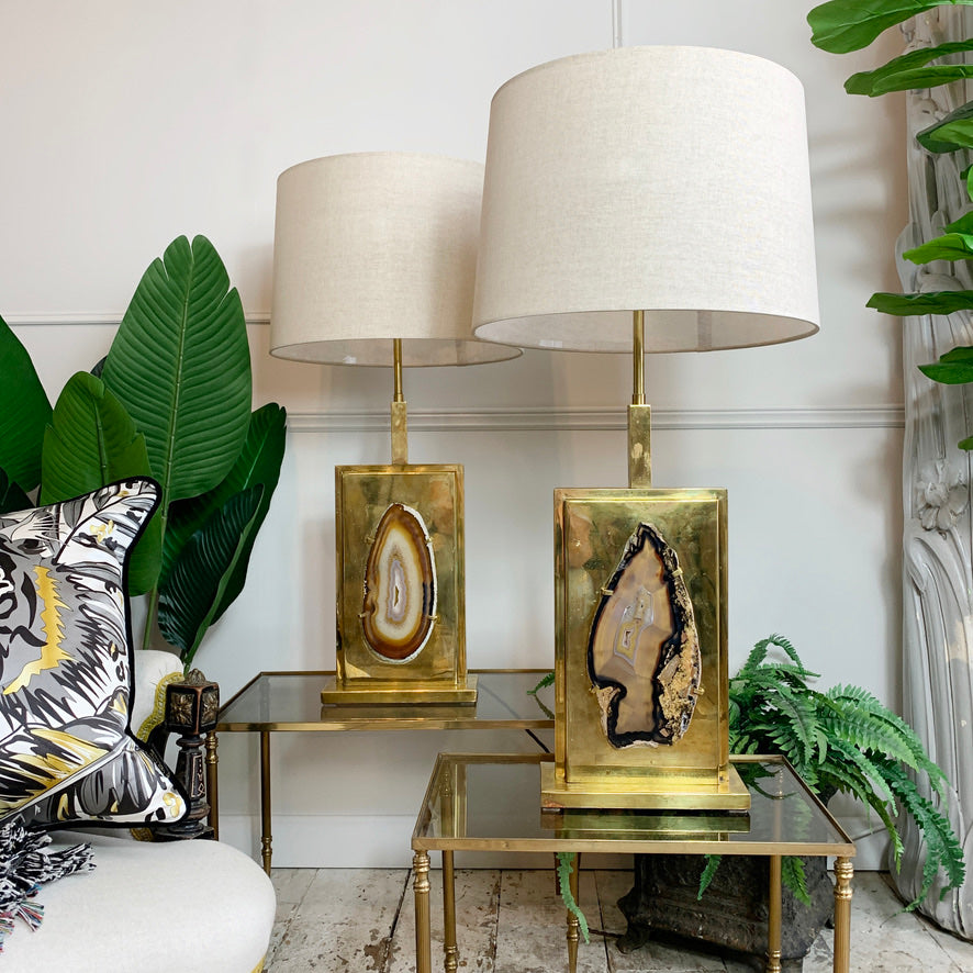 Two matching table lamps with large off-white drum shades and gold bases featuring geode slice inlays, styled on glass tables in a living room setting with plants and a sofa.