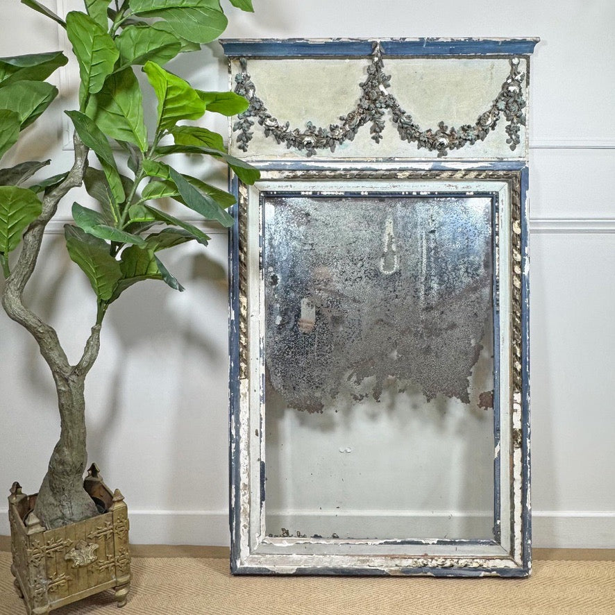 An antique French rectangular mirror with a distressed blue and white frame, leaning against a white wall next to a potted green plant.