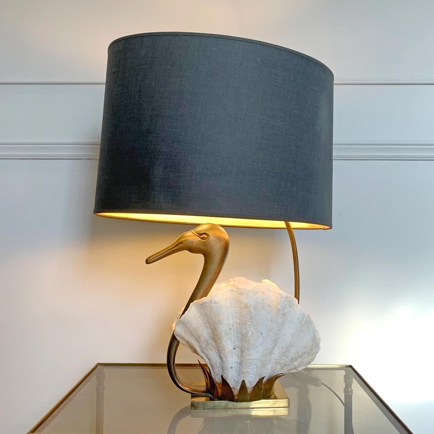A Willy Daro Swan table lamp with a large dark gray fabric drum shade, a gold-toned bird-shaped neck, and a white clam seashell base, shown lit on a glass tabletop against a white paneled wall.