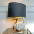 A Willy Daro Swan table lamp with a large dark gray fabric drum shade, a gold-toned bird-shaped neck, and a white clam seashell base, shown lit on a glass tabletop against a white paneled wall.