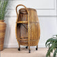 A tall, woven rattan or bamboo rolling storage cart with a curved handle, two open shelves, and black caster wheels, shown in an indoor room setting with a white paneled wall, beige carpet, and potted plants.