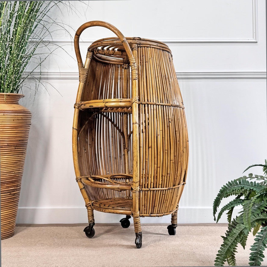 A tall, woven rattan or bamboo rolling storage cart with a curved handle, two open shelves, and black caster wheels, shown in an indoor room setting with a white paneled wall, beige carpet, and potted plants.