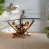 A decorative Maison Jansen console table with a clear oval glass top and a sculptural metallic base resembling leaves and branches, shown in a styled interior with a manetti pineapple ice bucket on top