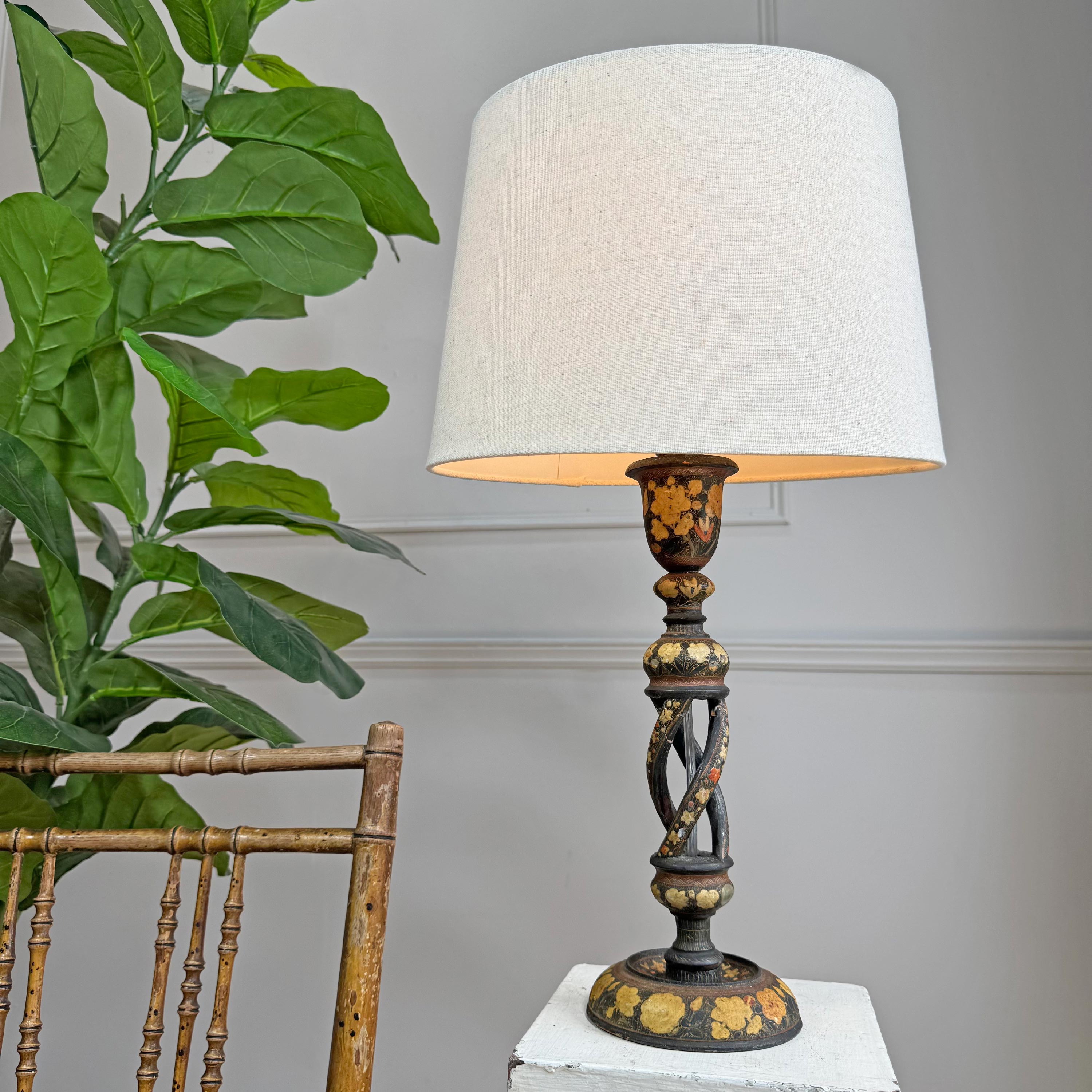 Decorative table lamp with a white shade next to a plant and wooden chair against a gray wall.