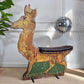 A vintage wooden reindeer fairground ride with a distressed gold and brown finish, a dark brown seat cushion, and green painted panels on the lower legs, set against a white panelled wall with a hanging disco ball.