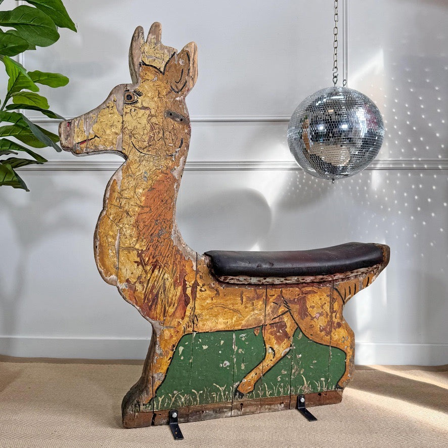 A vintage wooden reindeer fairground ride with a distressed gold and brown finish, a dark brown seat cushion, and green painted panels on the lower legs, set against a white panelled wall with a hanging disco ball.