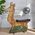 A vintage wooden reindeer fairground ride with a distressed gold and brown finish, a dark brown seat cushion, and green painted panels on the lower legs, set against a white panelled wall with a hanging disco ball.
