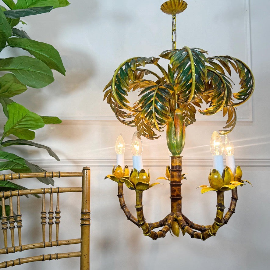 An antique vintage French palm tree-shaped chandelier with a metallic gold and green finish, featuring four candle-style lights, shown hanging in a room next to a bamboo chair and a green plant.