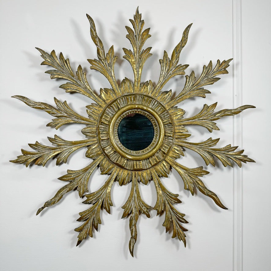 A decorative antique gold wooden sunburst wall mirror with ornate gold leaf-style leaf-shaped arms and a dark oval mirror at the center, mounted on a white wall.