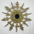 A decorative antique gold wooden sunburst wall mirror with ornate gold leaf-style leaf-shaped arms and a dark oval mirror at the center, mounted on a white wall.