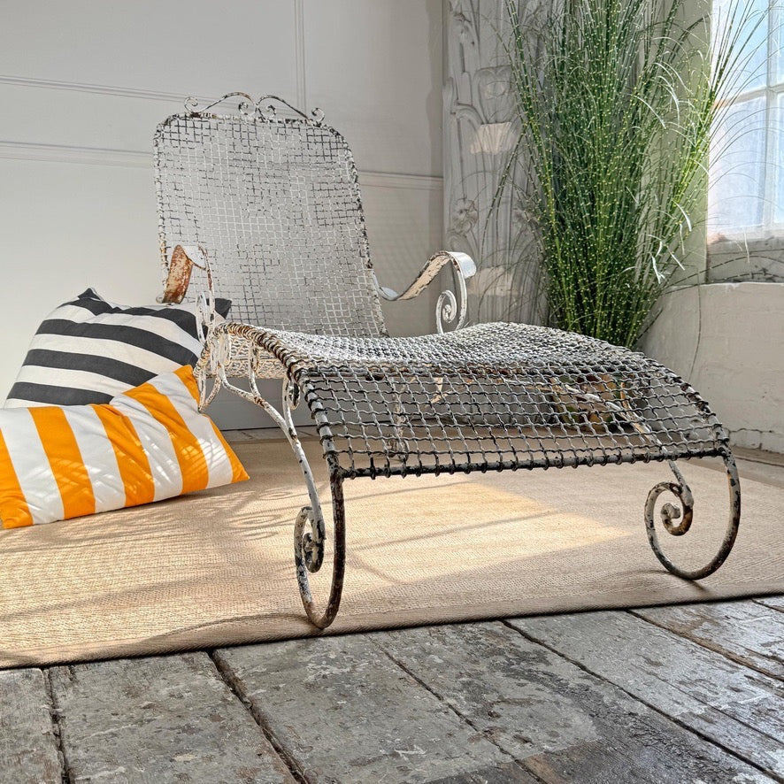 A rustic antique wirework metal wire chaise lounge chair with scrollwork legs, styled on a beige rug with striped and orange-and-white pillows against a white paneled wall.
