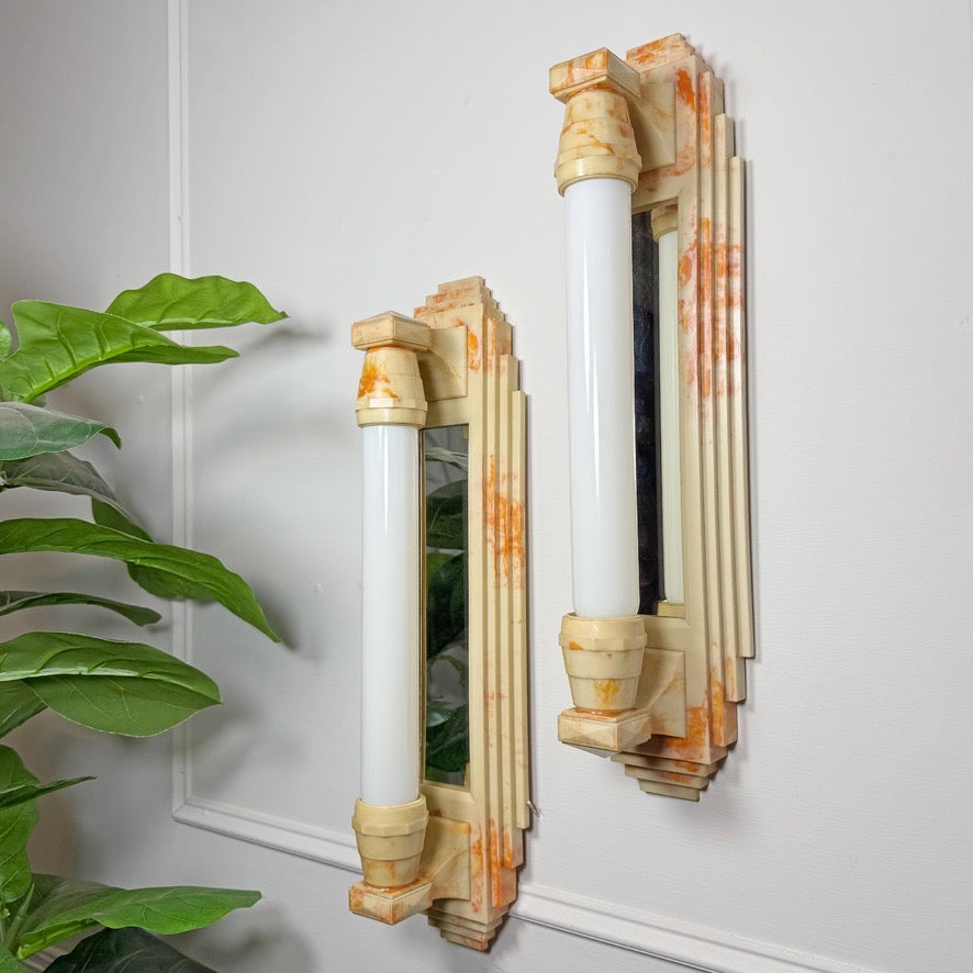 Two matching Art Deco wall sconces with cream and orange marbled bakelite frames and white cylindrical light bulbs, mounted on a white paneled wall with a green plant visible on the left.