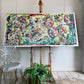 An abstract painting with layered graffiti-style markings in yellow, green, red, black, white, and blue, displayed on a wooden easel in a bright room with a white chair and a potted fern in the foreground.