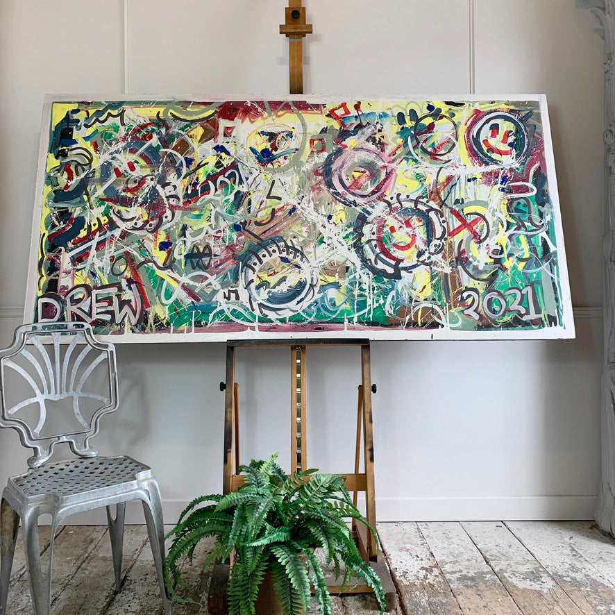 An abstract painting with layered graffiti-style markings in yellow, green, red, black, white, and blue, displayed on a wooden easel in a bright room with a white chair and a potted fern in the foreground.