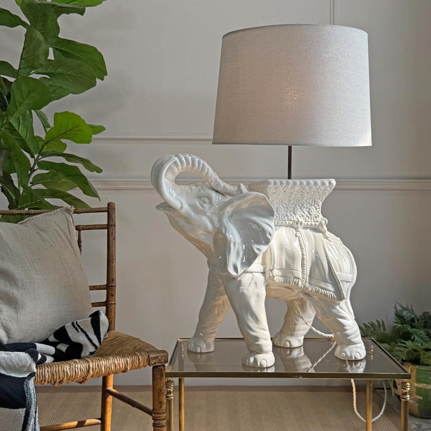 White ceramic elephanttable  lamp on a glass table with a chair and plant in the background