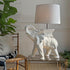 White ceramic elephanttable  lamp on a glass table with a chair and plant in the background