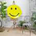 A large yellow smiley face sign mounted on a wooden easel, styled in a room with potted plants, a gray metal chair, and a white paneled wall.