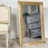 An ornate gold-framed antique floor mirror leaning against a white wall, reflecting a distressed gray wooden chest of drawers and a potted plant. Part of a matching upholstered chair is visible on the left.