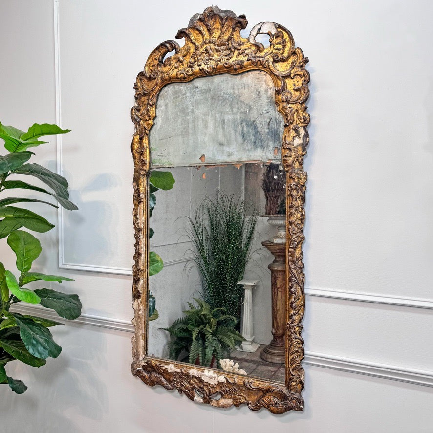 An ornate, Queen Anne split plate wall mirror with a heavily carved gold frame, mounted on a white paneled wall next to a green potted plant.