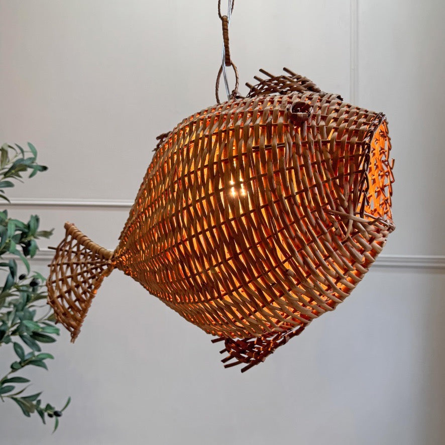 A fish-shaped woven pendant light made from natural brown reed or wicker, hanging against a white wall with a small green plant visible on the left.