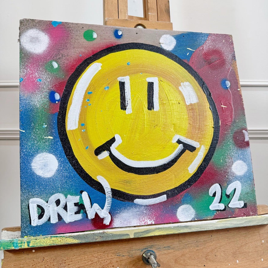 outsider artist Drew, A square hand-painted canvas featuring a large yellow smiley face with black and white details, set against a colorful abstract background with paint splatters. The canvas is displayed on a wooden easel, with the name 'DREW' and the number '22' painted in white at the bottom.