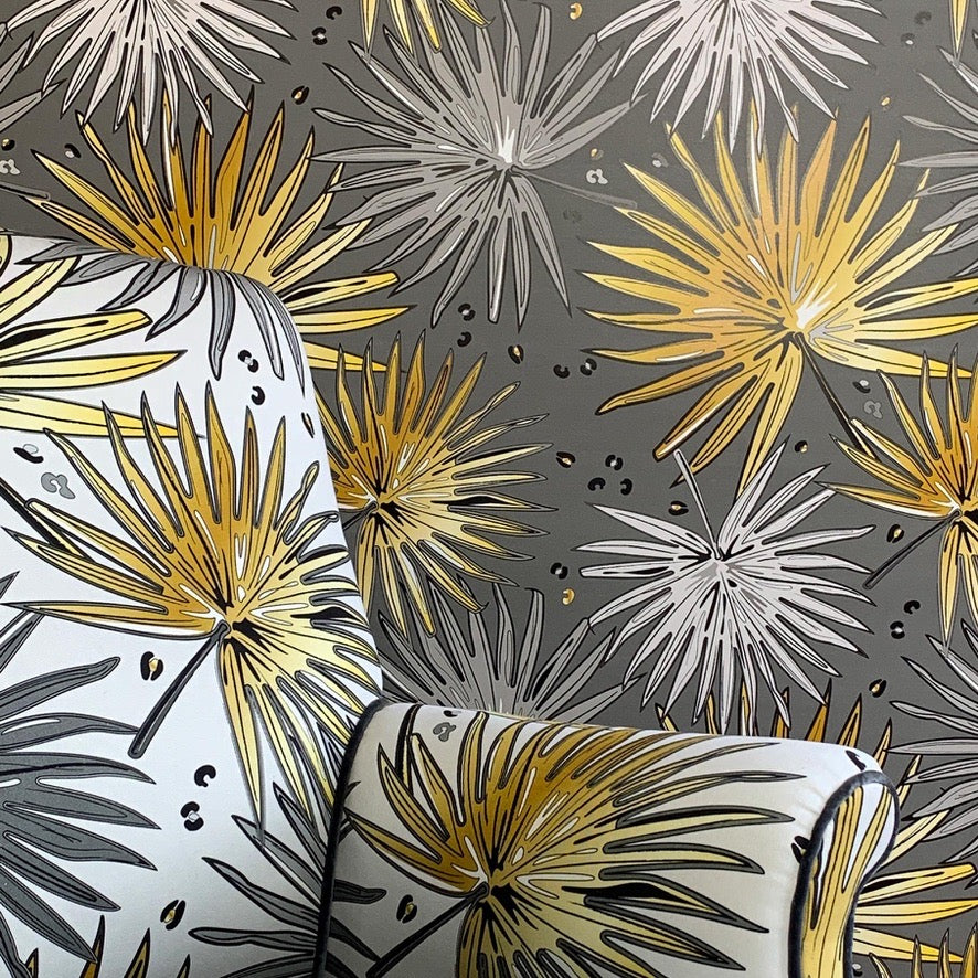 A close-up of a patterned fabric or wallpaper featuring stylized palm fronds in metallic gold, white, and dark gray on a dark gray background.