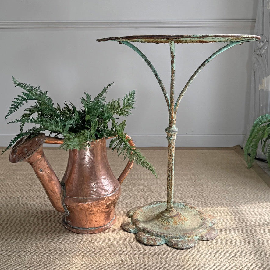 A rustic copper watering can filled with green ferns beside a distressed metal table with a green patina finish, set on a burlap surface against a neutral wall.