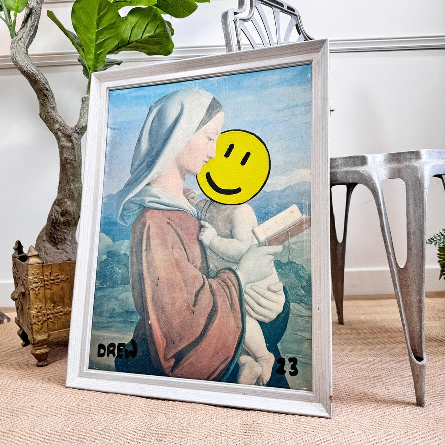 A framed art print featuring a painting of the Virgin Mary holding the infant Jesus, styled in a home setting with a potted tree and a metal chair in the background. The face area of the print is obscured by a yellow smiley face graphic.