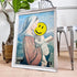 A framed art print featuring a painting of the Virgin Mary holding the infant Jesus, styled in a home setting with a potted tree and a metal chair in the background. The face area of the print is obscured by a yellow smiley face graphic.