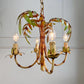 A rustic palm tree themed chandelier with faux bamboo stalks, green leaves, and four candle-style bulbs with clear glass shades, shown hanging against a light gray paneled wall.
