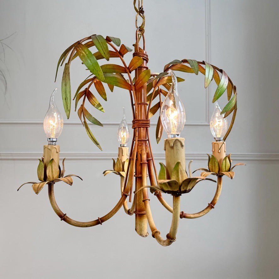 A rustic palm tree themed chandelier with faux bamboo stalks, green leaves, and four candle-style bulbs with clear glass shades, shown hanging against a light gray paneled wall.