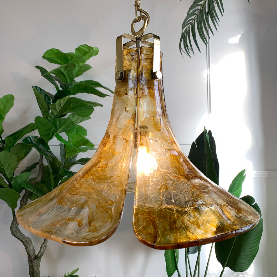 A vintage pendant light with curved amber-tinted lucite shades, hanging against a white wall with green houseplants visible in the background.