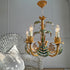 A decorative vintage faux bamboo palm  chandelier with a gold-toned metal frame and green painted palm leaves, shown hanging in a styled interior next to a white wire chair.