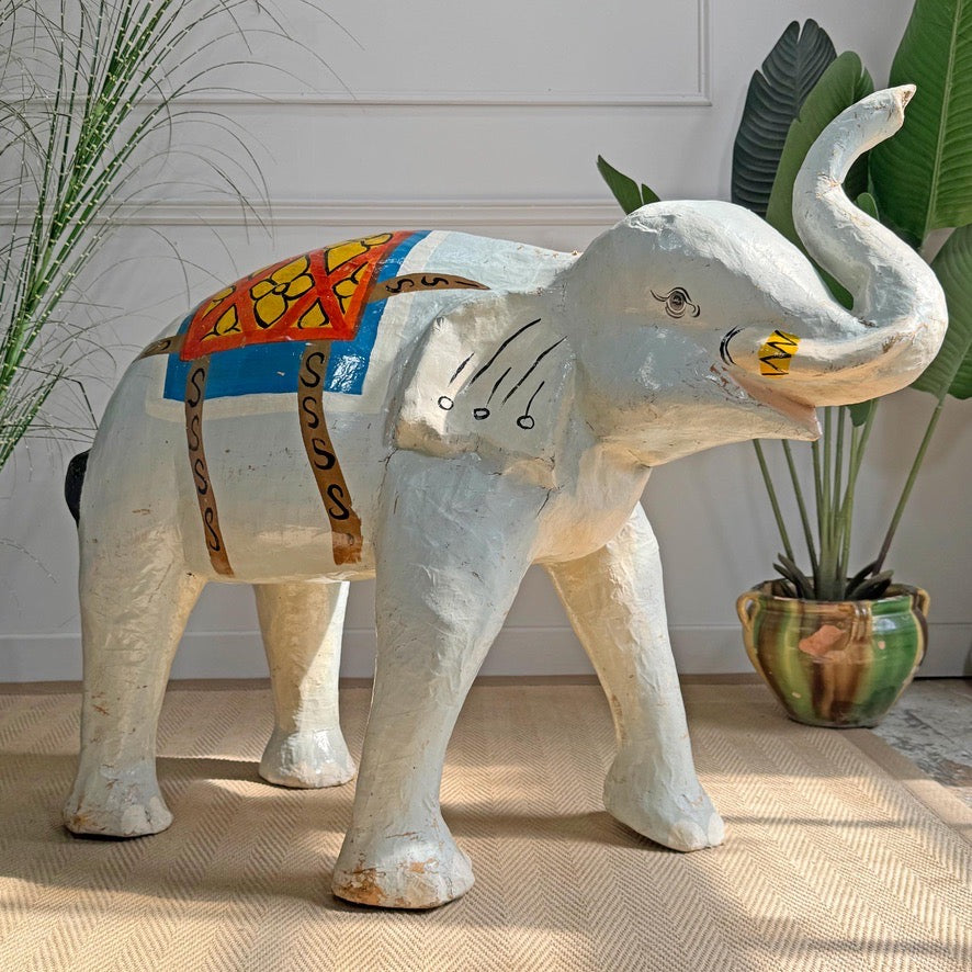 A hand-painted vintage grey paper mache elephant statue with a colorful painted saddle and decorative details, standing on a textured beige rug in a styled indoor setting with potted plants.