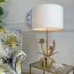 A maison jansen gold flower table lamp with a large white drum shade and a sculptural gold metal base featuring leaf and flower motifs, placed on a glass-topped side table next to a patterned armchair in a styled interior setting.