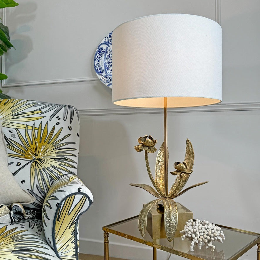 A maison jansen gold flower table lamp with a large white drum shade and a sculptural gold metal base featuring leaf and flower motifs, placed on a glass-topped side table next to a patterned armchair in a styled interior setting.