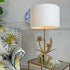 A maison jansen gold flower table lamp with a large white drum shade and a sculptural gold metal base featuring leaf and flower motifs, placed on a glass-topped side table next to a patterned armchair in a styled interior setting.