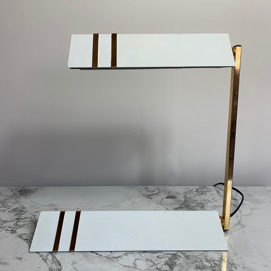 A vintage two-tier table lamp with white rectangular metal body featuring two gold stripes, supported by a gold metal frame, set on a marble surface against a gray wall.