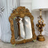 An ornate Late 19th Century Italian Carved Mirror with a baroque-style carved frame, leaning against a white decorative vase on a stone surface next to a gold floral table lamp.