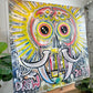 A colorful graffiti-style painting of a skull with large eyes and curved tusks, by Drew outsider artist, shown hanging on a wall in an indoor setting. The artwork includes the text 'DREW' and '2025' and is photographed against a white wall with plants and furniture partially visible.