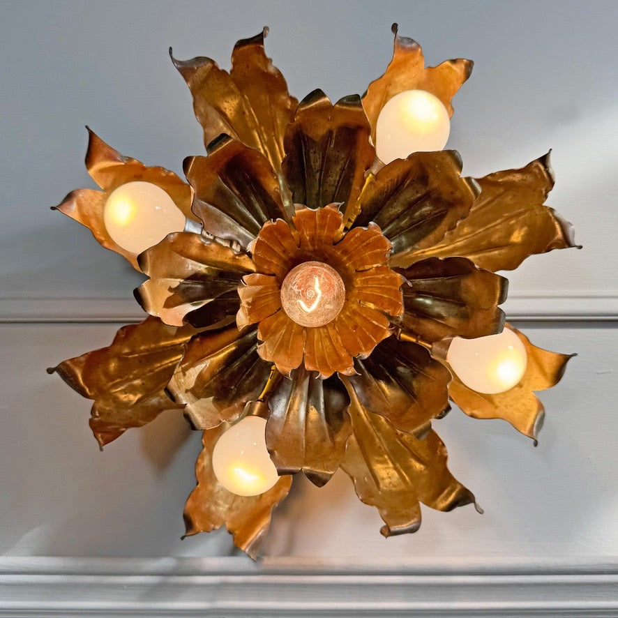 A decorative vintage brass and copper wall sconce shaped like a metallic flower with multiple leaf-like petals and six glowing frosted globe bulbs, mounted against a light gray wall.