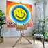 A colorful acrylic painting on an easel, by Drew outsider artist, featuring a yellow smiley face surrounded by concentric circles in blue, white, orange, and red, with the text &