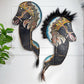 Two handcrafted Kuda Lumping horse head wall hangings displayed side-by-side on a white paneled wall, with a green plant leaf visible on the left.