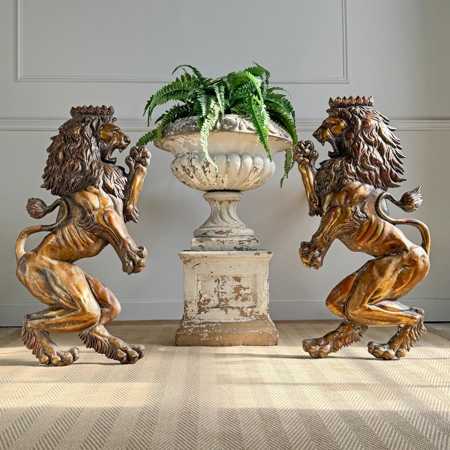 A pair of vintage ornate bronze lion plaques facing each other on either side of a distressed white stone urn pedestal with a green fern arrangement, set against a paneled gray wall and patterned beige carpet.